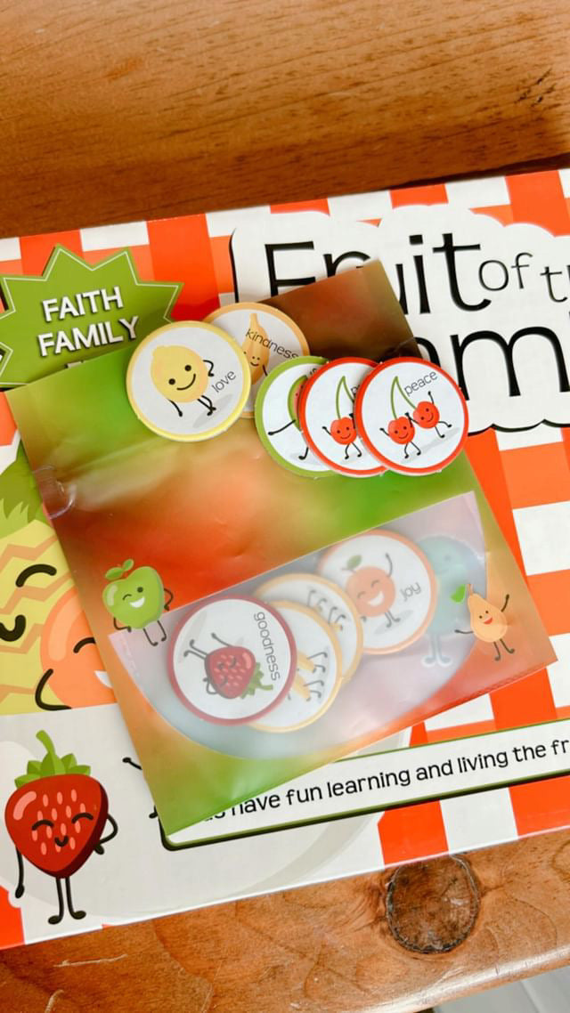 Closeup of a red and white checkered game box with words Fruit of the Family on it sitting on a wood table. Smiling strawberry in the lower left corner. On top of box is a zipper bag that looks like a fruit bowl. Tokens with pictures of fruit on them are in the bowl and on laying on top of the bag.