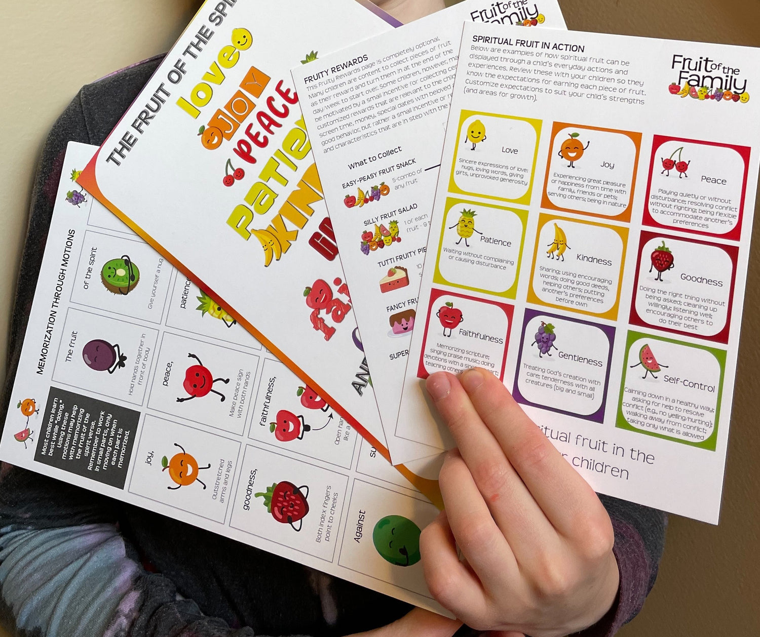 Close up of a hand holding 4 cards that are part of the game. Card one shows smiling fruit doing motions for a bible verse. Card two shows colorful fruit of the spirit bible verse. Card 3 is titled Fruity Rewards and card 4 is titled Spiritual Fruit in Action and has 9 boxes-each with a piece of fruit describing one of the fruits of the spirit.