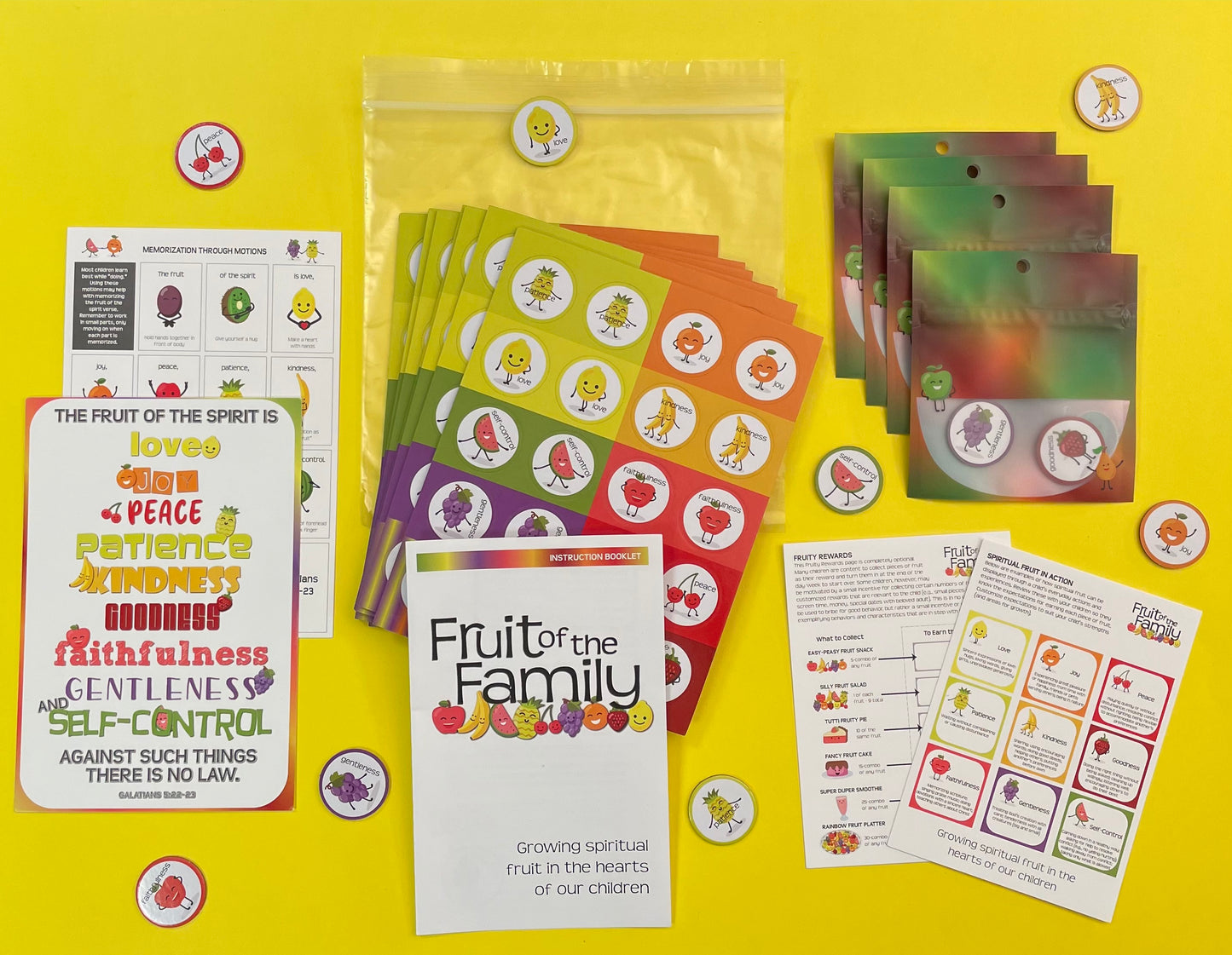 Bible verse card with the fruit of the spirit verse is displayed on a yellow background along with other game supplies, 9 boards of round tokens to be popped out, 4 bags that look like fruit bowls and tokens with pictures of fruit are scattered about.