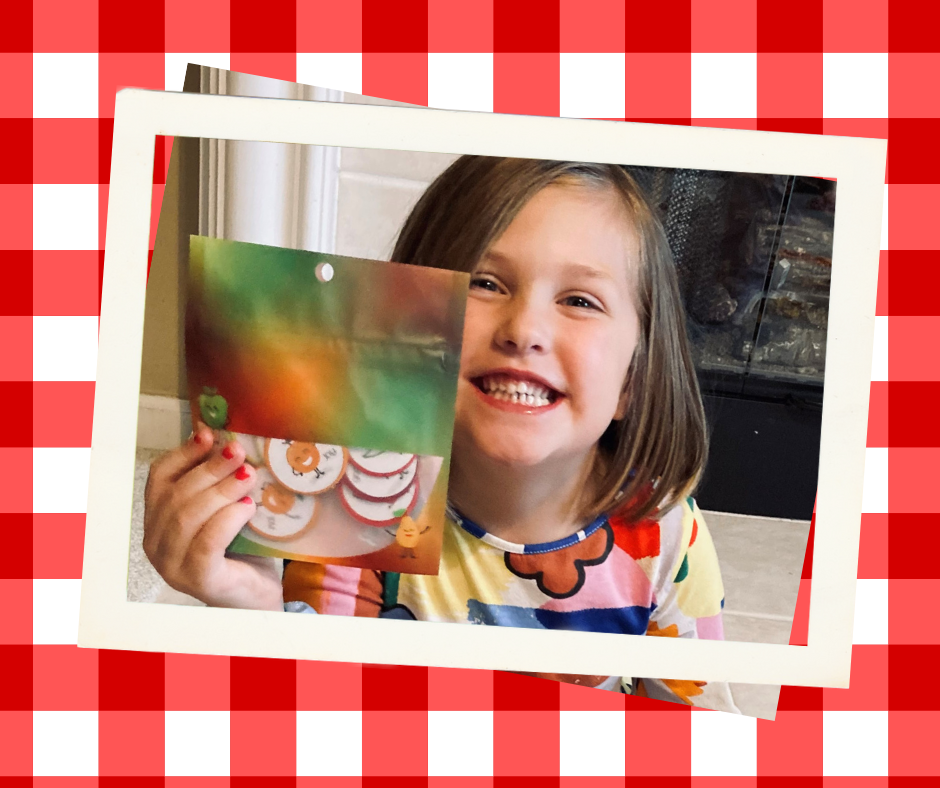 Close-up of a little girls face. She is smiling and holding a bag with the image of a fruit bowl on it. In the bag is a large group of tokens with pictures of smiling fruit on them.