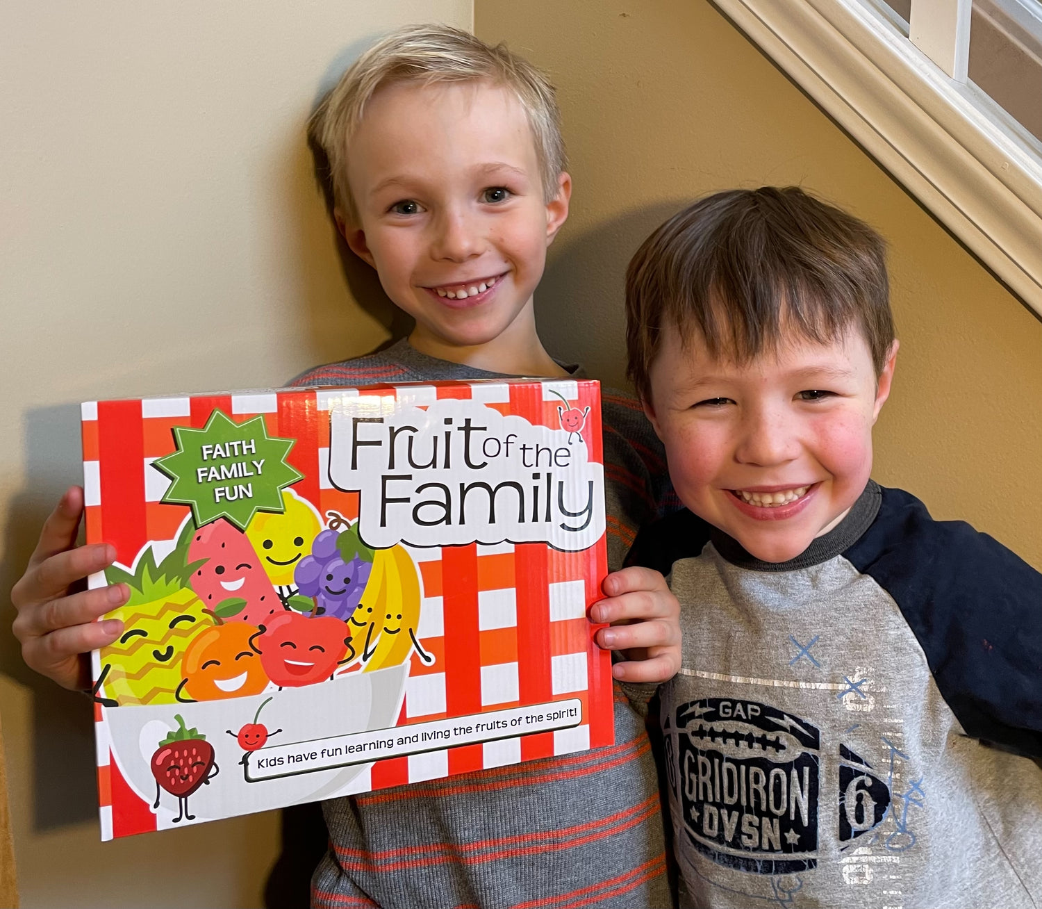 Two little boys standing side by side. One boy is holding a red and white checkered box with the name Fruit of the Family on it. The box also has a bowl full of smiling fruit with the words "faith, family, fun" above it. 