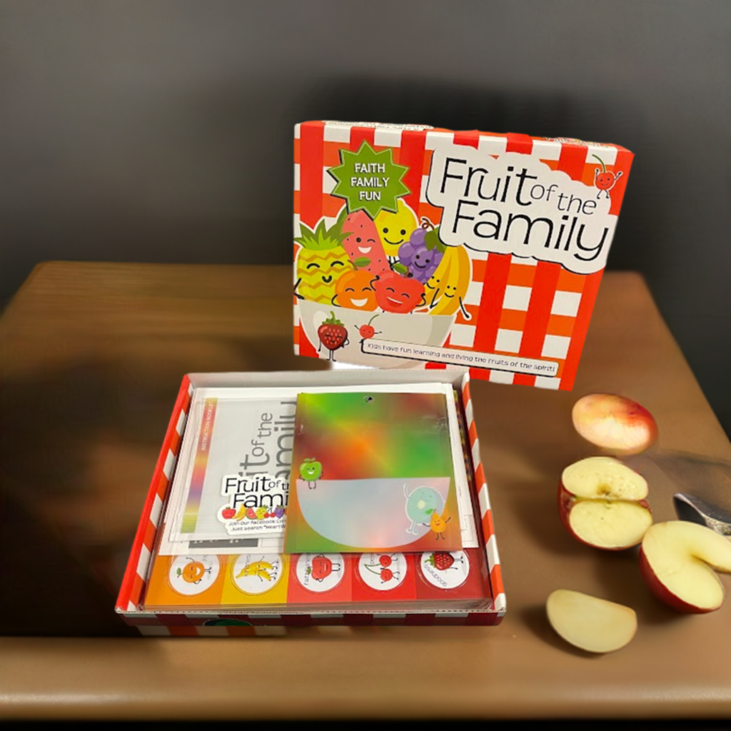 Red and white checkered game box is open and placed on a wooden table. A cut up apple is right next to it. The box top displays a bowl of smiling fruit with the words "faith, family, fun" above it. The name of the game, Fruit of the Family is on the box top. The contents of the box are visible in the open bottom portion of the box. 