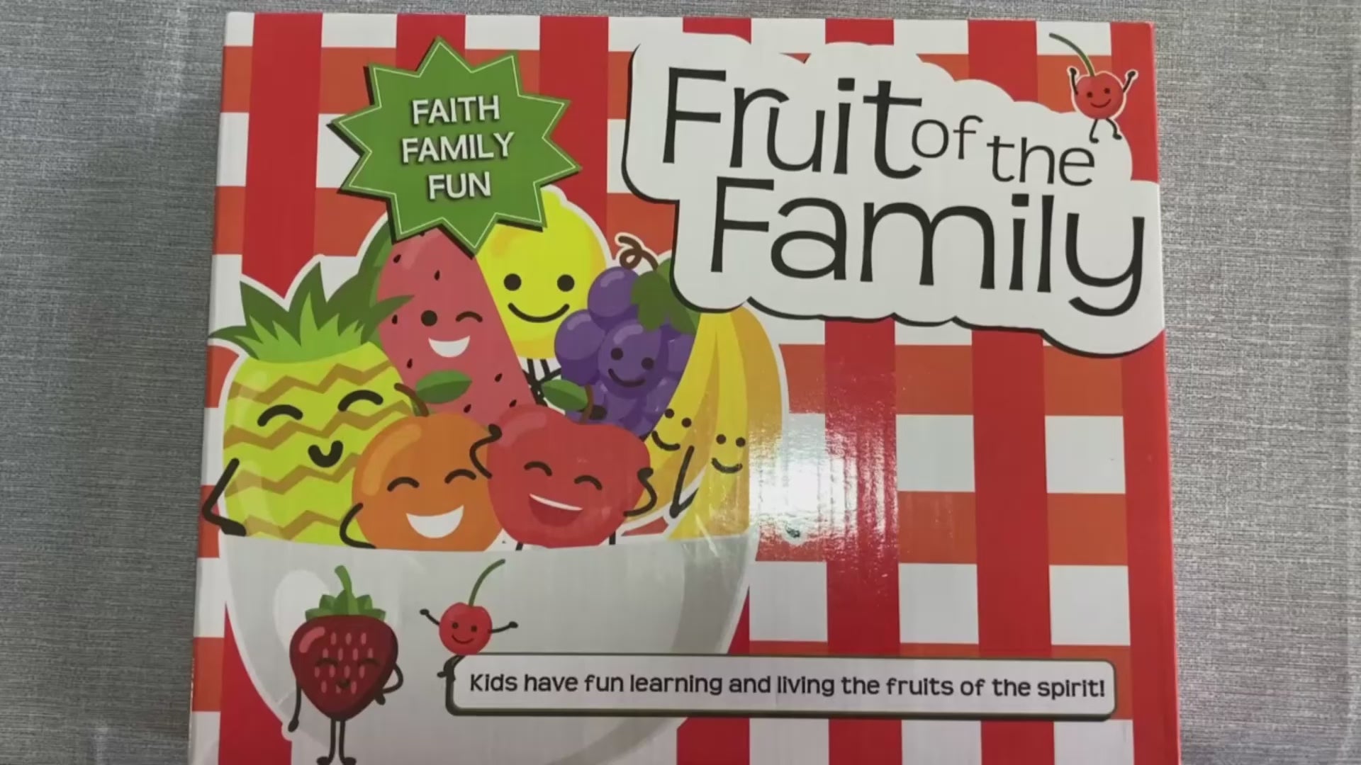 Load video: Video shows close-up of two hands opening a red and white checkered box with the title Fruit of the Family on the top. The box top has a bowl of smiling fruit with the words faith, family fun above it. The video shows the hands removing each piece of the game and showing the camera. Game pieces include 4 white cards with text, instruction booklet, 4 zippered bags with pictures of fruit bowl on them, and 9 boards of tokens with pictures of fruit on them to be popped out. 